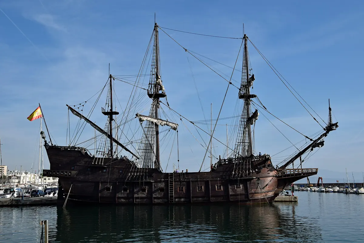A replica galleon with three tall masts and a Spanish flag at the stern, moored in a calm harbour